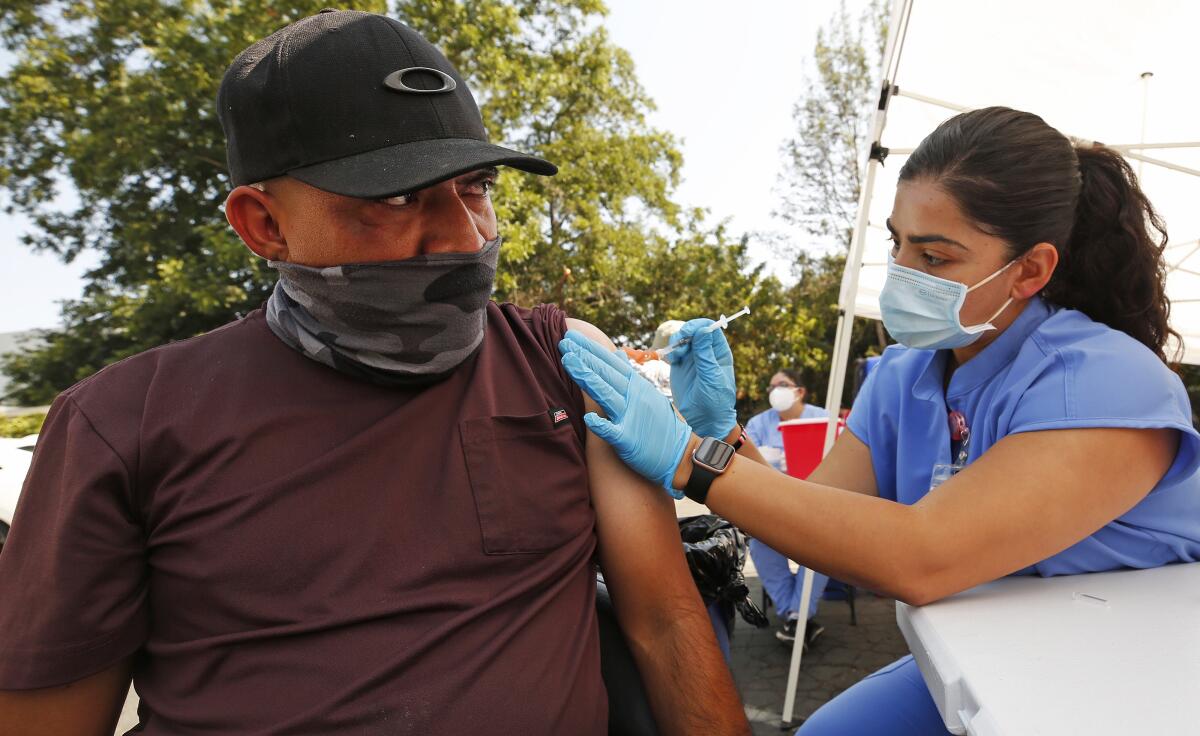 A man in a mask sits as a woman in protective gear administers a shot.