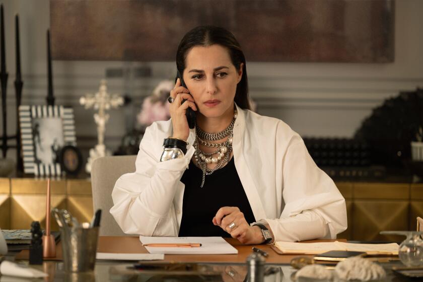 A woman in heavy jewelry sits at a desk talking on the phone.