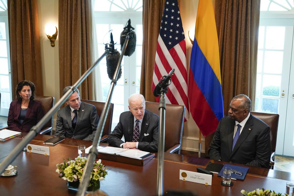 Microphones loom over a woman, from left, and three men seated at a table near two flags 