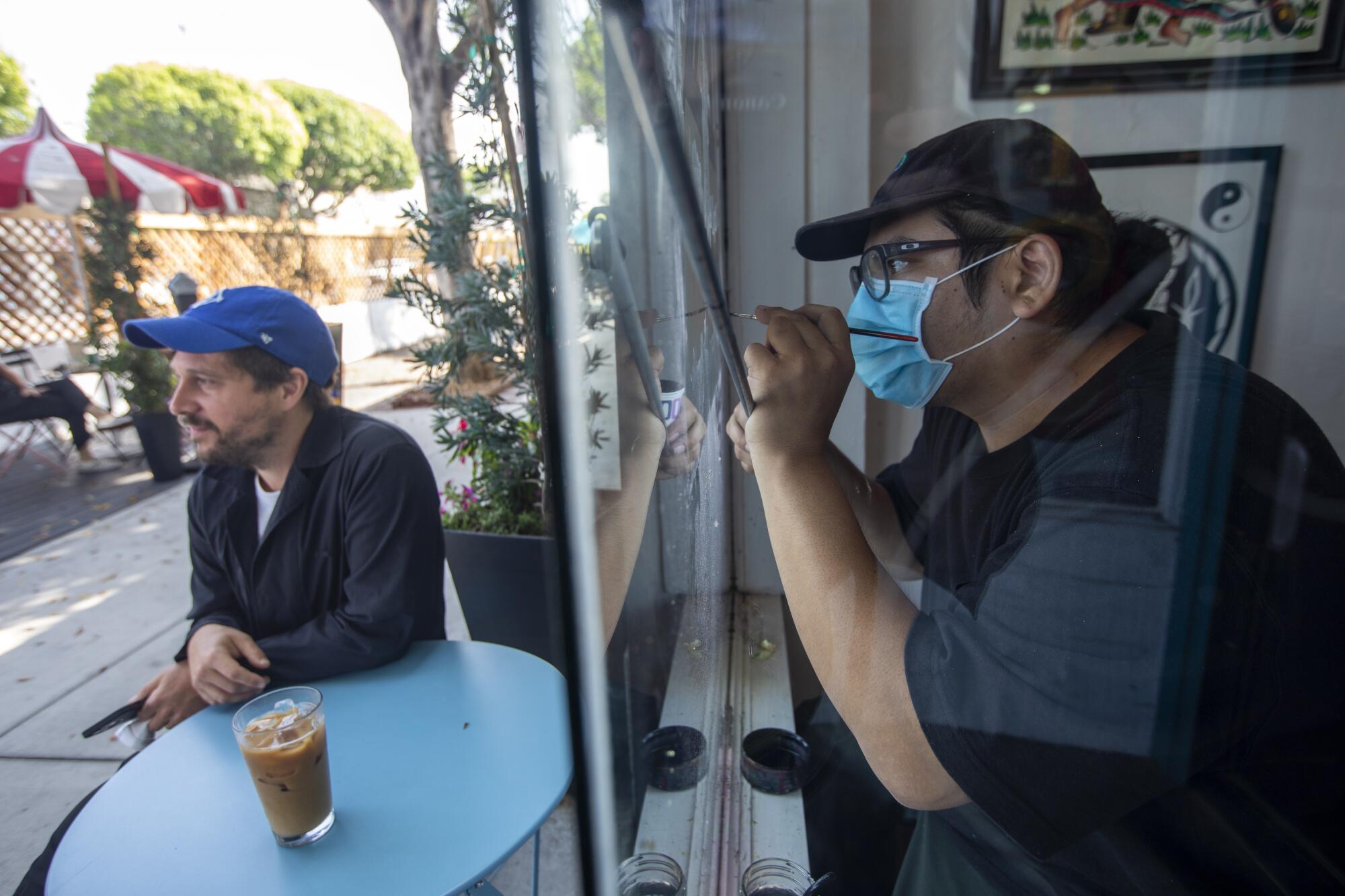 An unmasked man sits at an outdoor table with an iced coffee as another man wearing a mask paints the inside of a shop window
