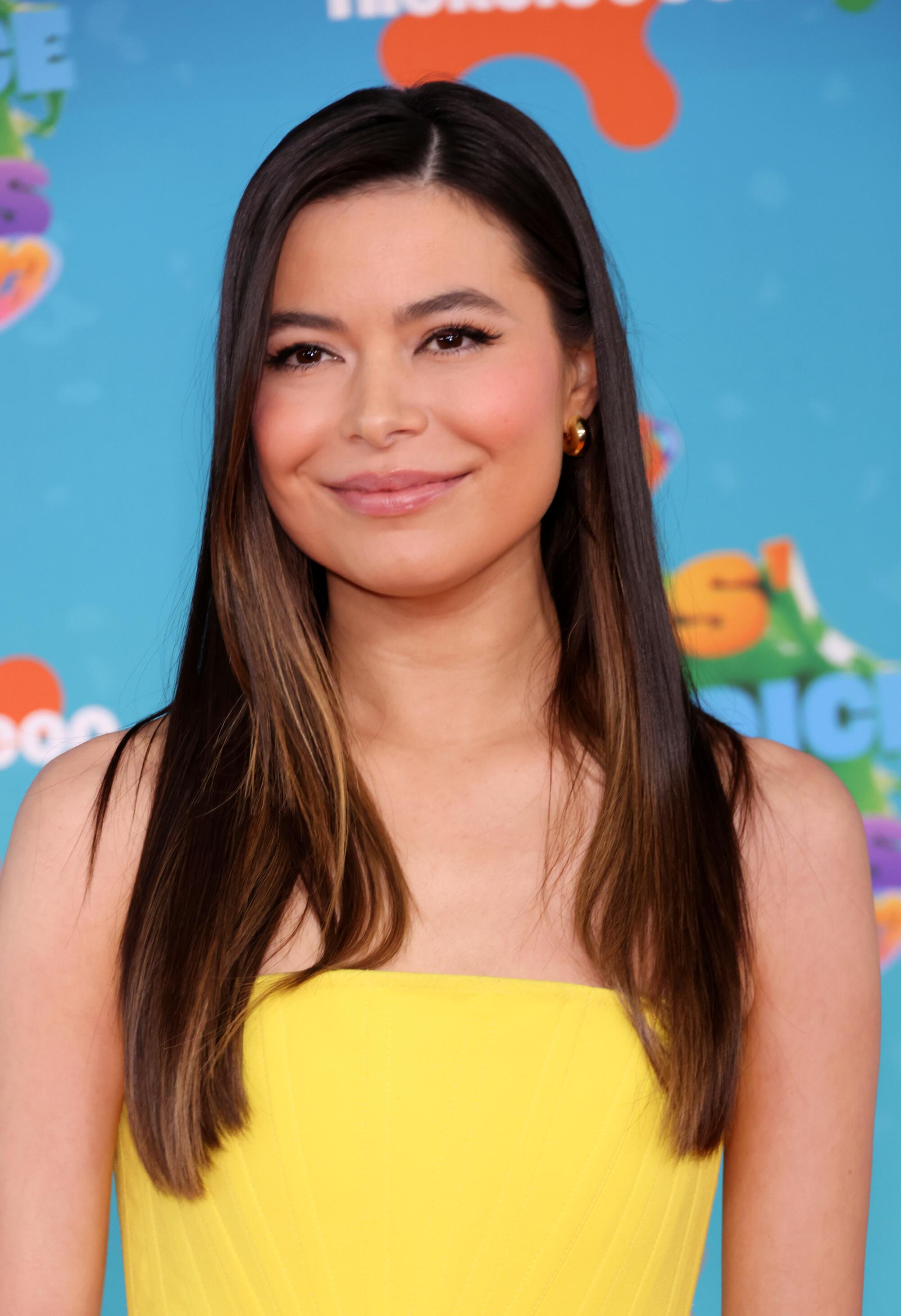 Miranda Cosgrove, wearing a yellow strapless dress, poses in front of a blue Nickelodeon backdrop. 