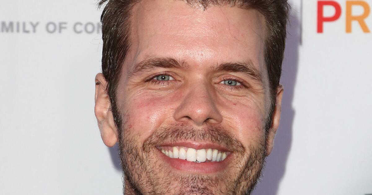Perez Hilton smiles in a camo jacket and poses.