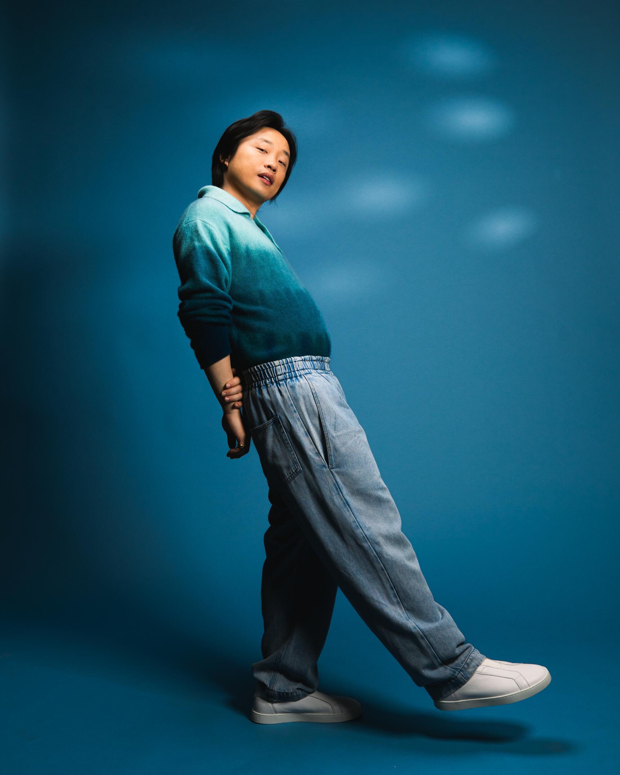 Jimmy O. Yang in a blue ombre sweater, pants and white shoes standing in front of a blue backdrop.