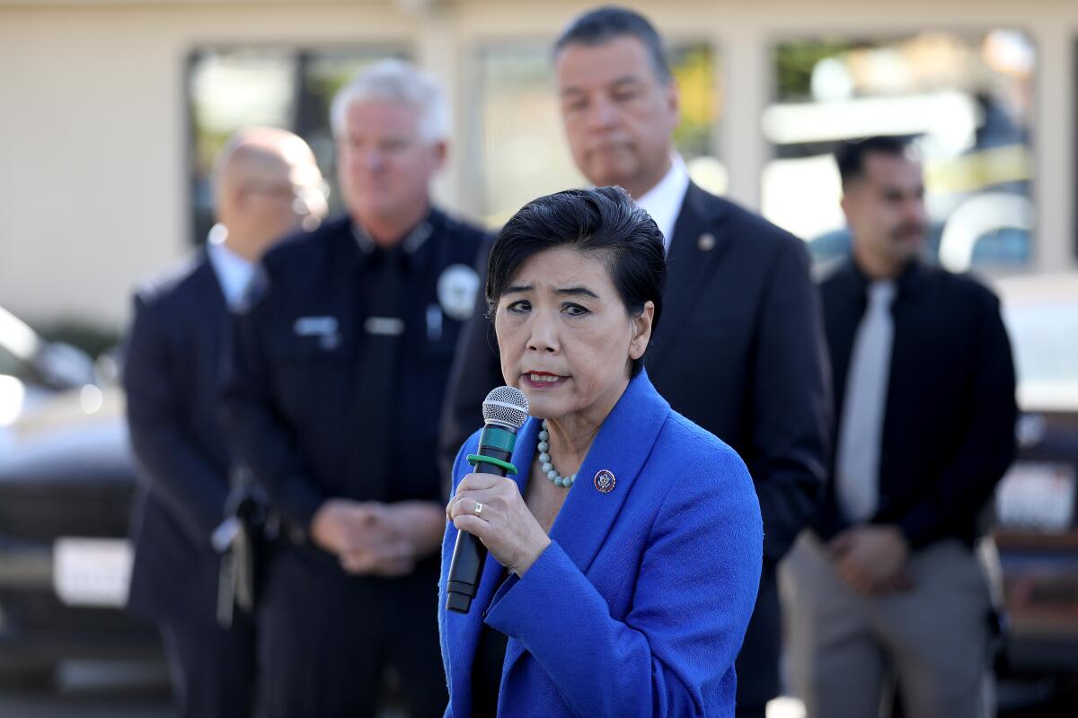 A woman with short dark hair and a blue suit jacket speaks into a microphone.