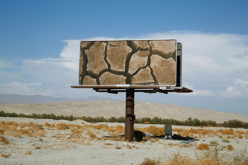 A billboard in the middle of a desert landscape features a photograph of a dry, cracked lakebed