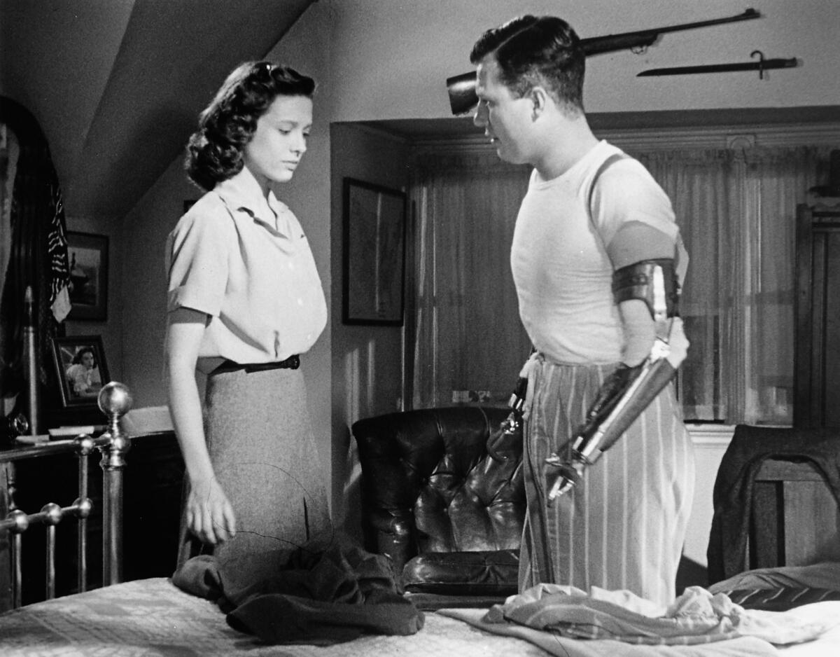 A woman and a man who has artificial arms and hands stand talking, facing each other, in a black-and-white movie still