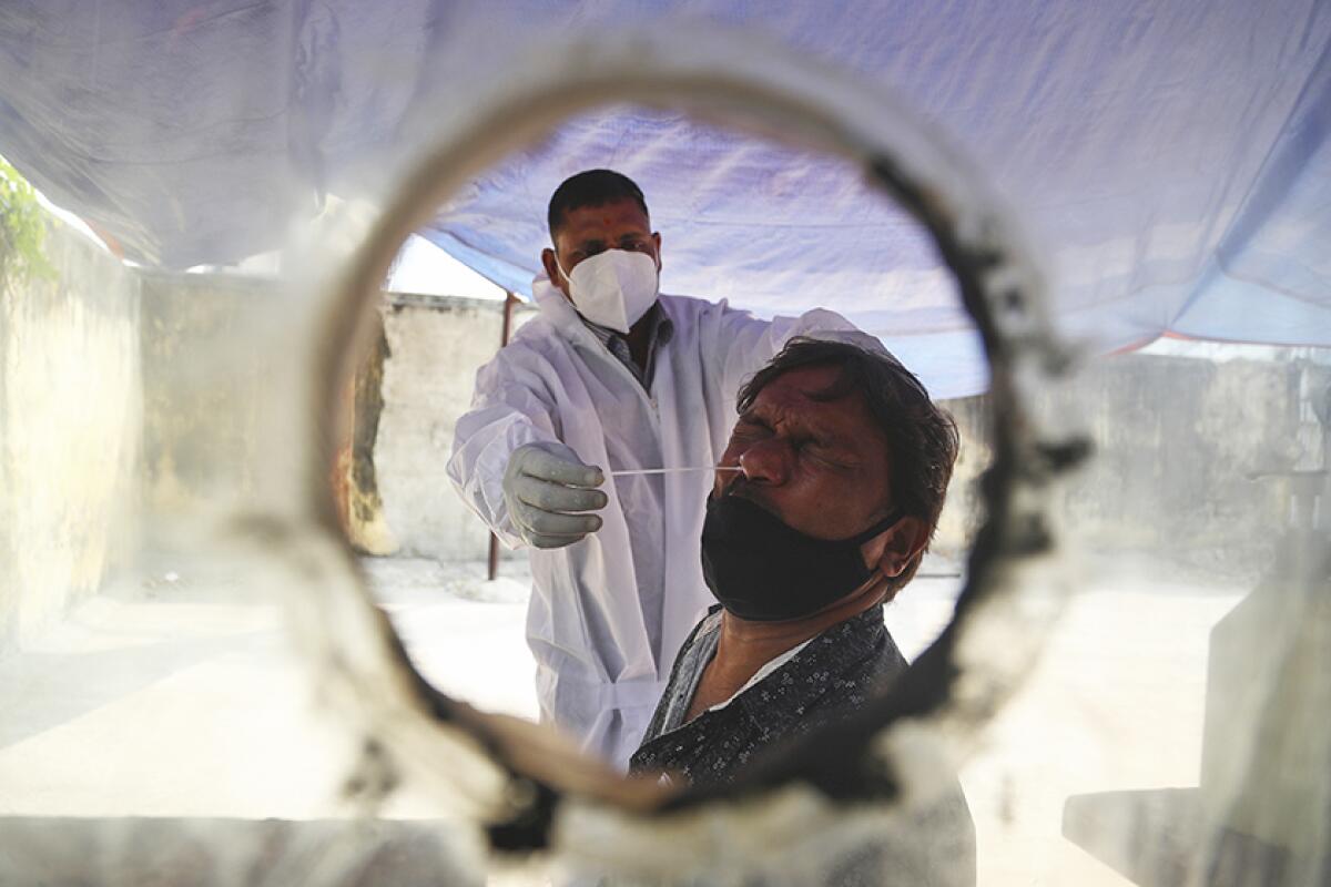 A man in protective gear inserts a nasal swab into another man's nose, seen through a circular hole in plastic sheeting.