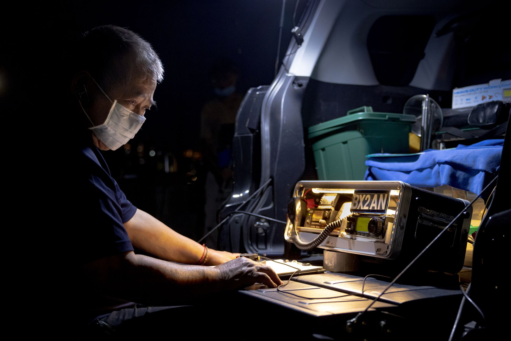A masked man seated outside the open trunk of a van looks at a machine with the word "BX2AN" on the top right corner.