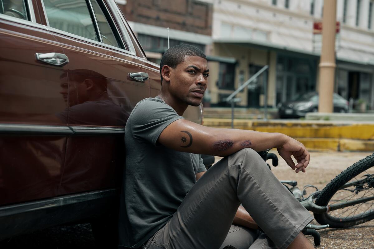 A man sits on the ground, leaning against a car, a bike on the ground next to him