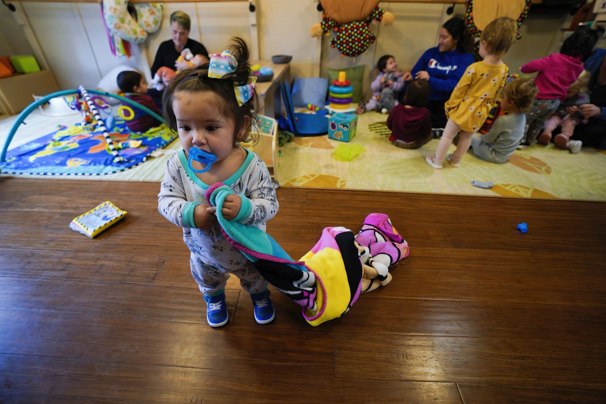 A toddler stands next to a toy, sucking a pacifier and holding a blanket, as women care for other children in the background.