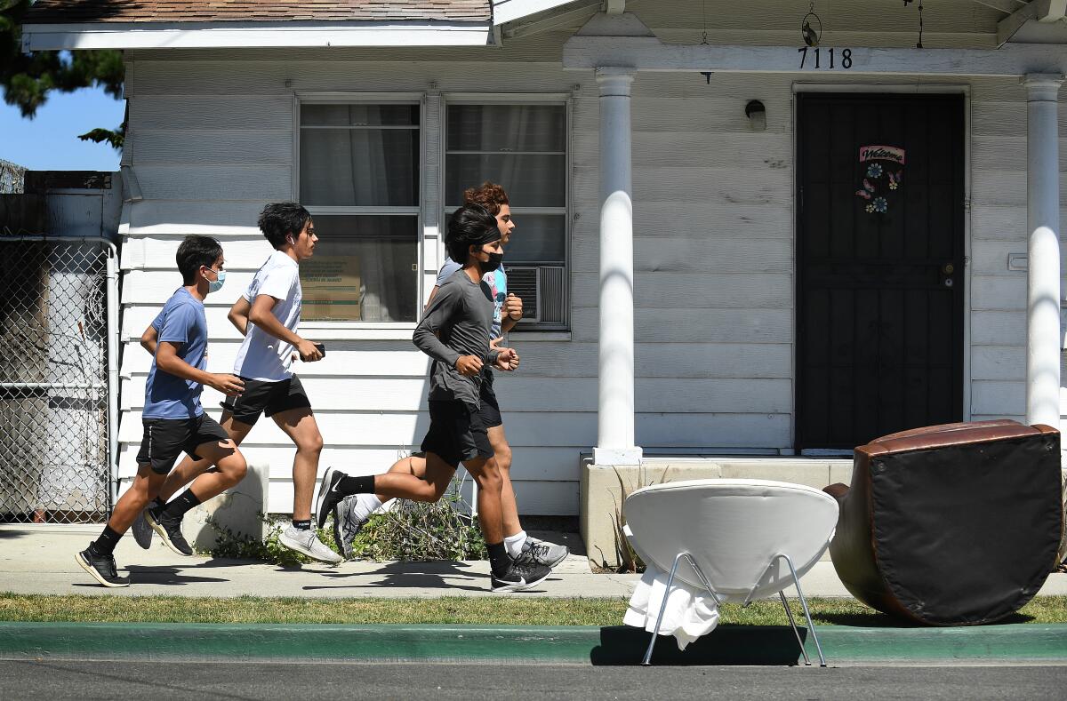 Four people, two of them in masks, jog along a city sidewalk past the front of a small house.