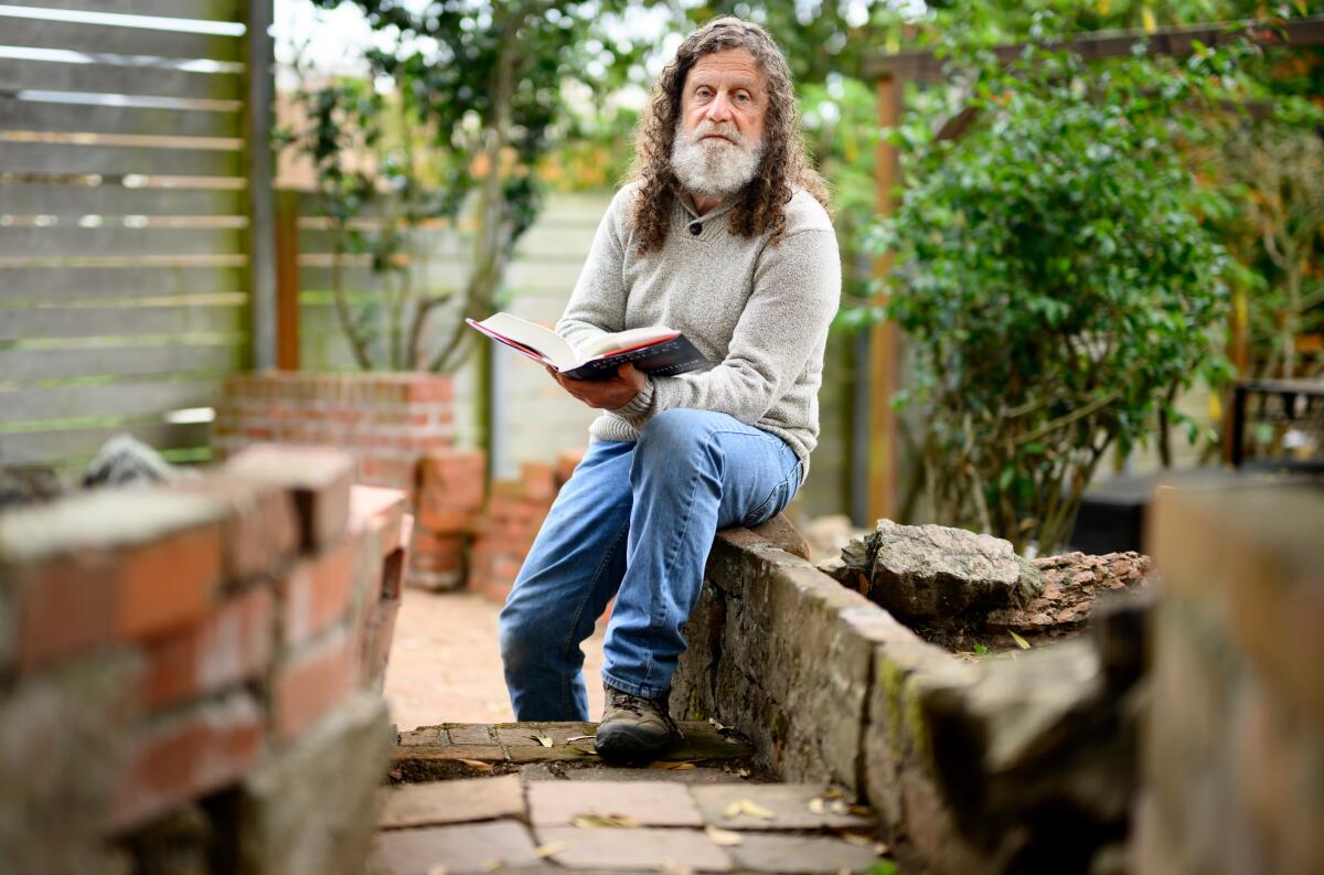 A man with curly brown hair and gray beard sits holding an open book, with plants behind him