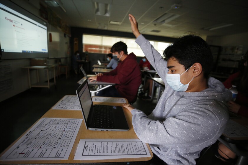 A masked student sitting at a desk with a laptop in a classroom raises his hand.
