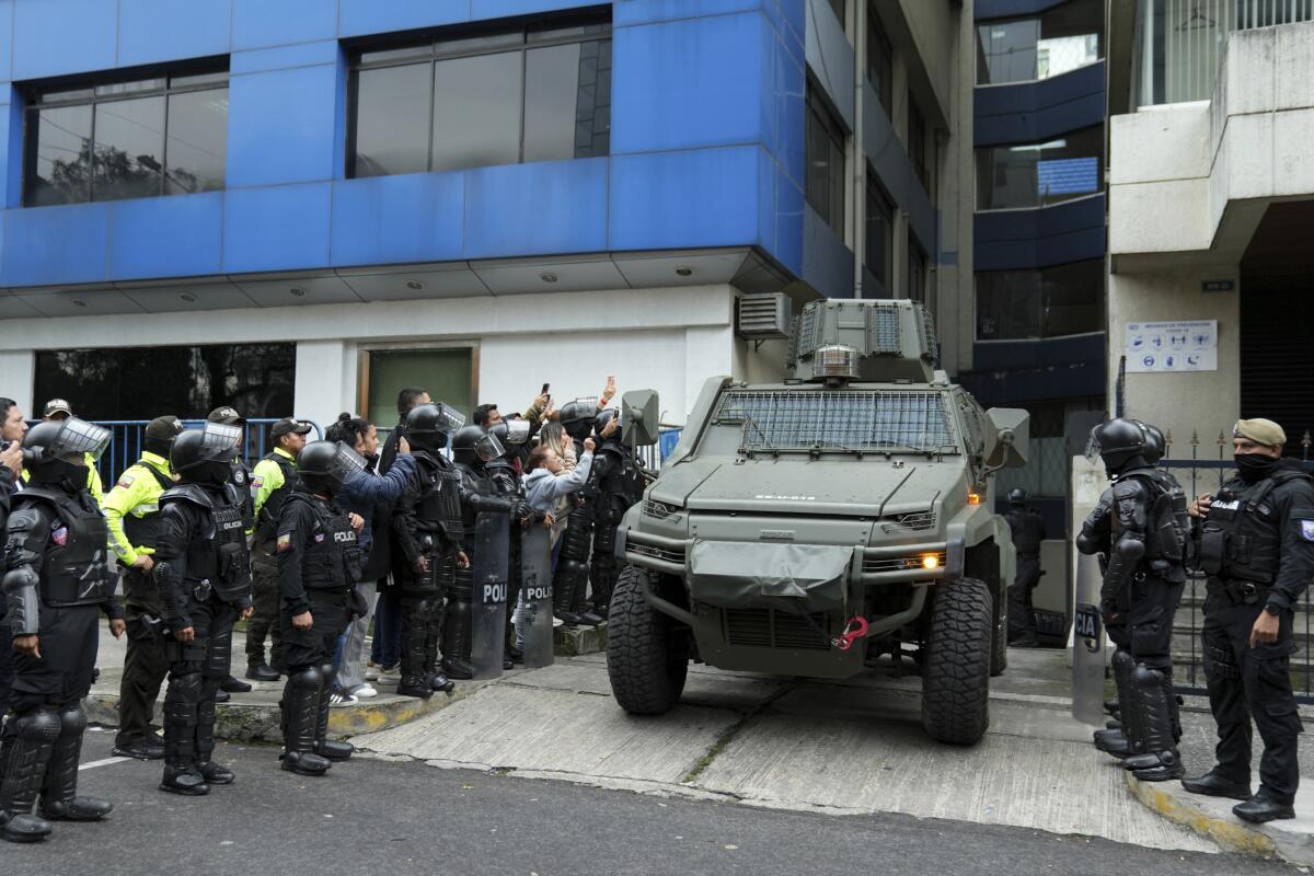 A military vehicle leaves a detention center as people in helmets and body armor stand on either side.