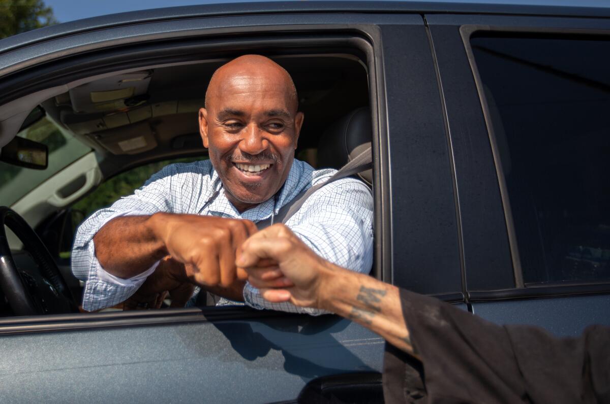 A smiling man seated in a driver's seat of a car exchanges a fist bump