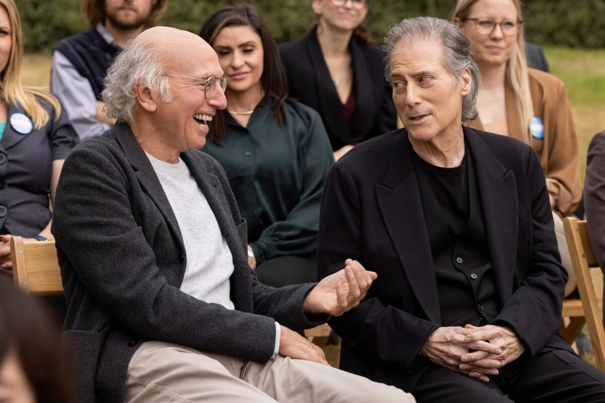 Larry David and Richard Lewis laugh and talk while sitting at a casual event with others behind them