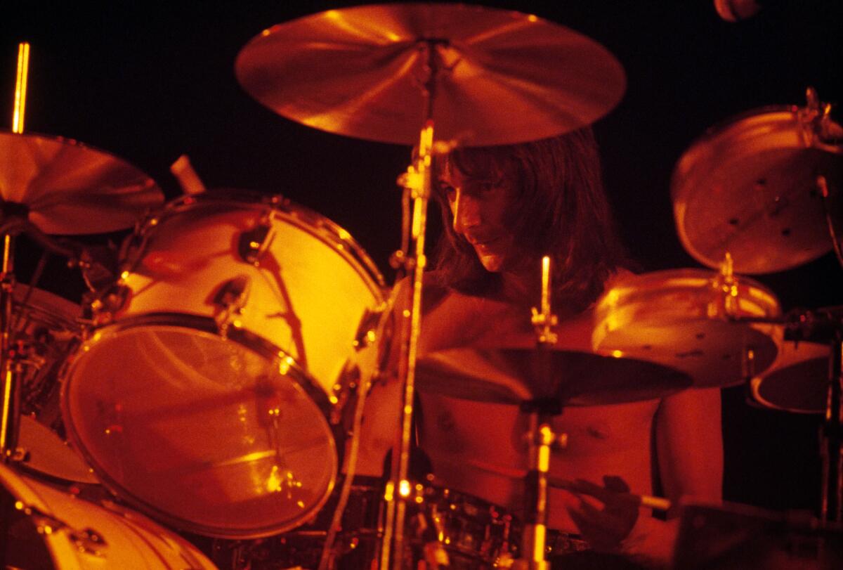 A man with long hair and no shirt can barely be seen drumming behind a drum set lighted in orange tones