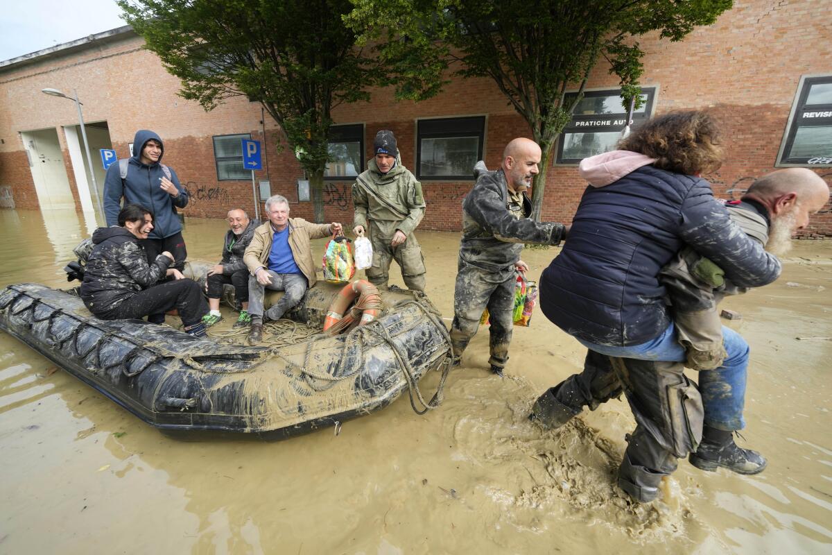 A man standing amid muddy waters carries a woman on his back. Nearby are people seated in an inflatable boat 