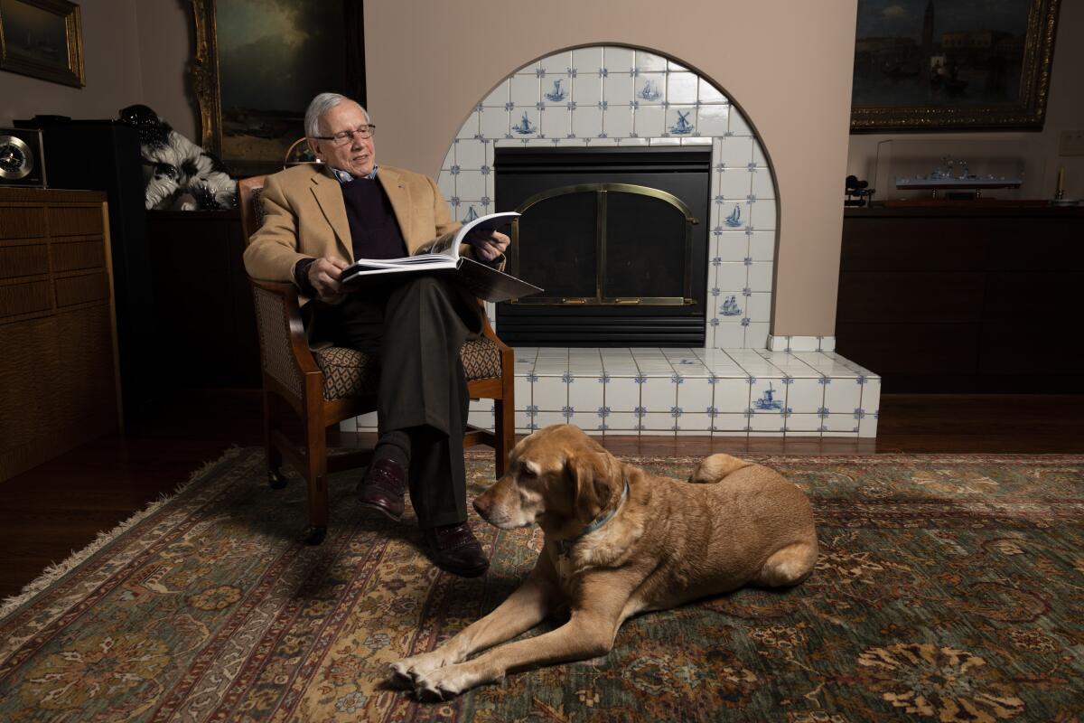 A man, seated, looks through a book. His dog lies in front of him.
