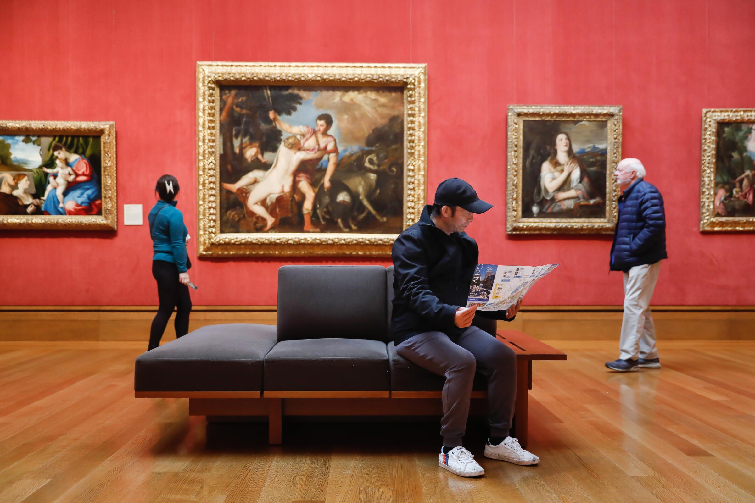 A young man reads a program while sitting on a museum bench in a gallery with red walls hung with Renaissance paintings.