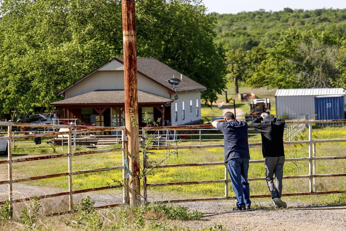 Two men lean against a fence looking away and toward a rural house surrounded by lush greenery.