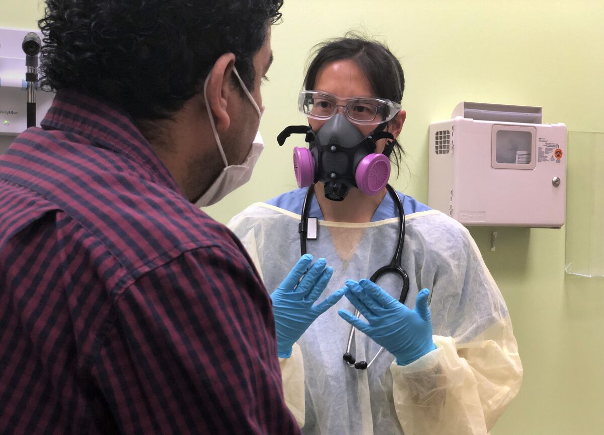 A healthcare worker in goggles and a mask speaks to a man in an exam room