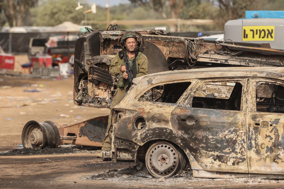 A man in military uniform stands next to the blackened remains of a car 