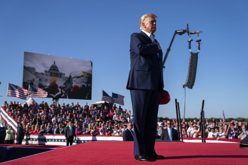 FILE - As footage from the Jan. 6, 2021, insurrection at the U.S. Capitol is displayed in the background, former President Donald Trump stands while a song, "Justice for All," is played during a campaign rally at Waco Regional Airport, Saturday, March 25, 2023, in Waco, Texas. The song features a choir of men imprisoned for their role in the Jan. 6, 2021, insurrection singing the national anthem and a recording of Trump reciting the Pledge of Allegiance. (AP Photo/Evan Vucci, File)