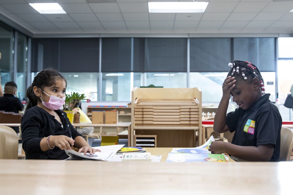 Two little girls, one in a pink mask drooping below her nose, sit facing each other at a table reading picture books.