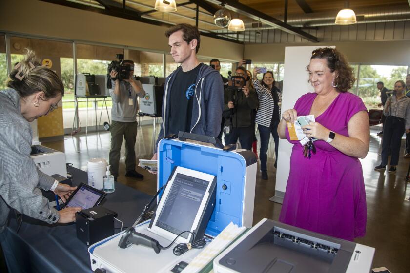 Irvine, CA - March 02: Rep. Katie Porter, right, and her son, Luke Hoffman, center, 18, who is a first-time voter, is helped by voter center customer service representative Jennifer Martin at a voting station at University Hills Community Center Saturday, March 2, 2024 in Irvine, CA. (Ringo Chiu / For The Los Angeles Times)
