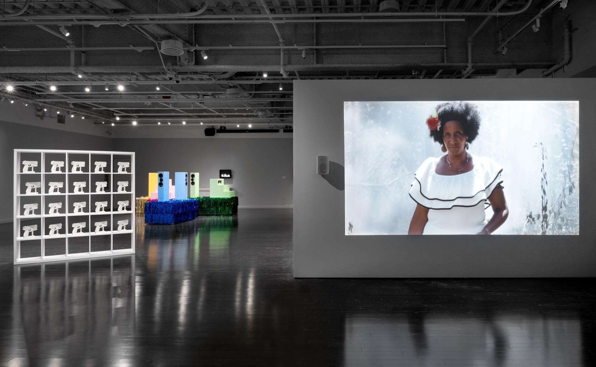 An art gallery contains two sculptural installations — one all in white, one in color — as well as a video projection