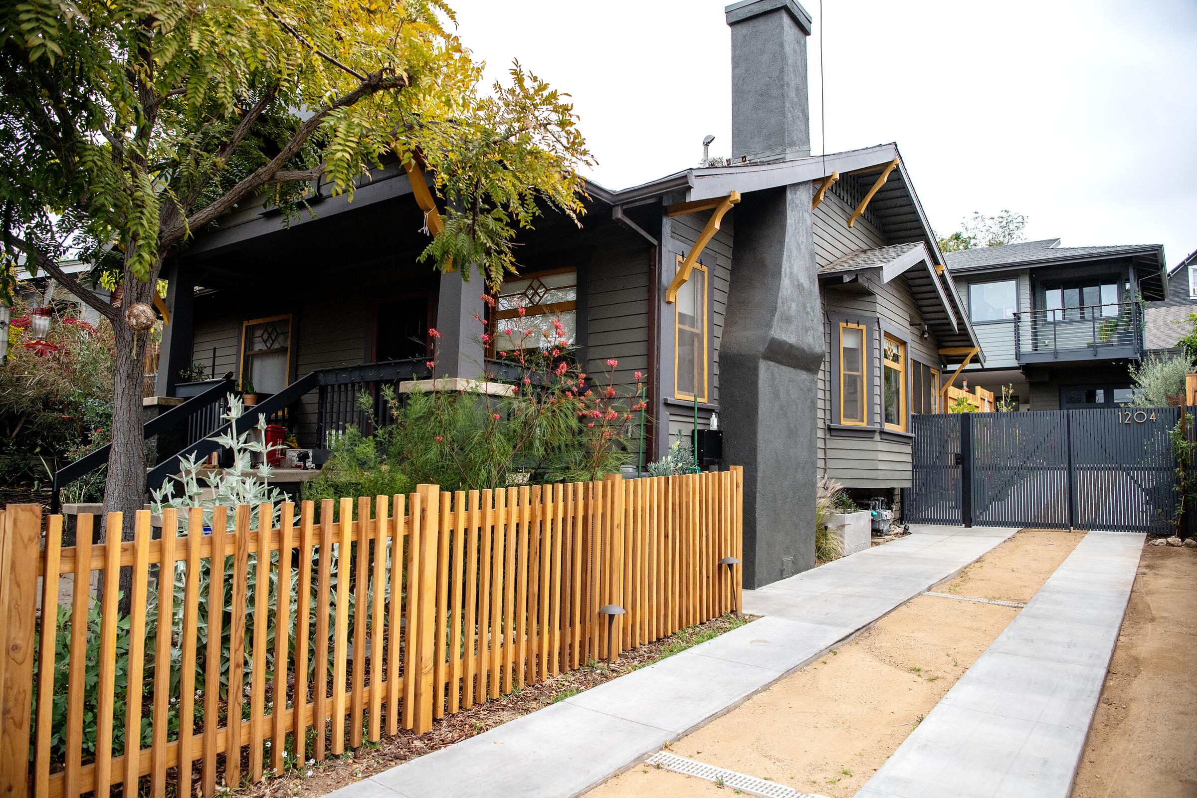 A gray house with yellow trim has a wood picket fence in the front yard and an accessory dwelling unit elevated behind it. 