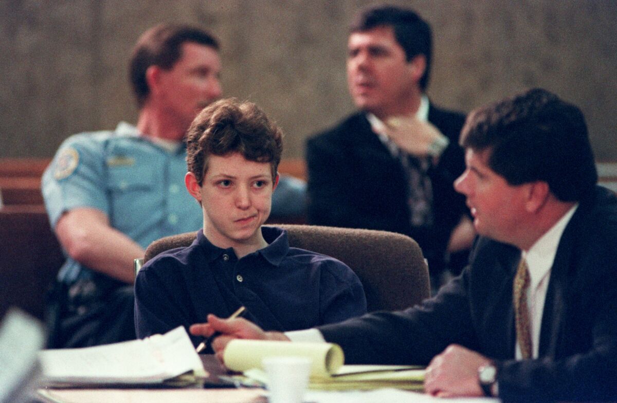 A young man sits in a courtroom with an attorney, with two men in the background.