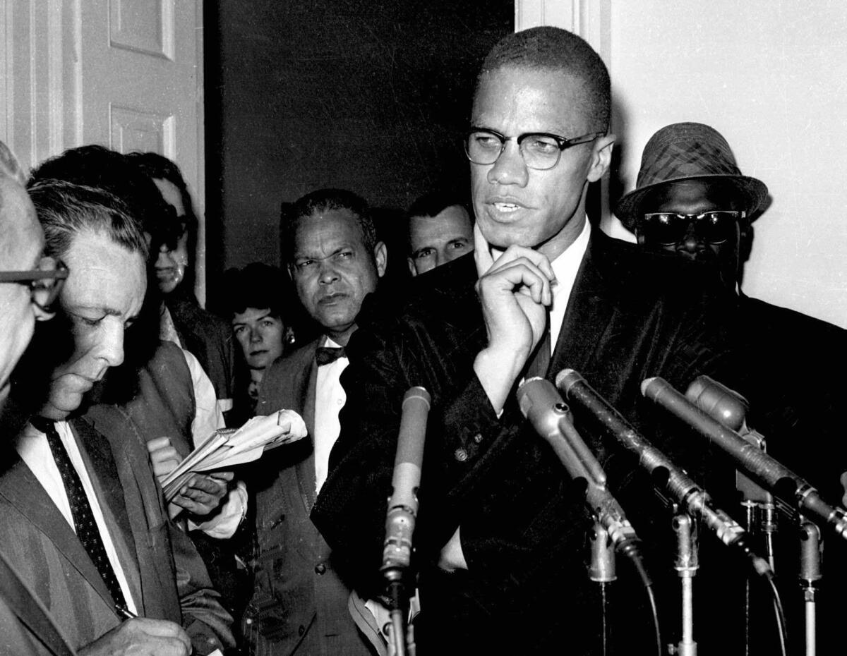 A tall man wearing glasses speaks to a crowd of reporters.