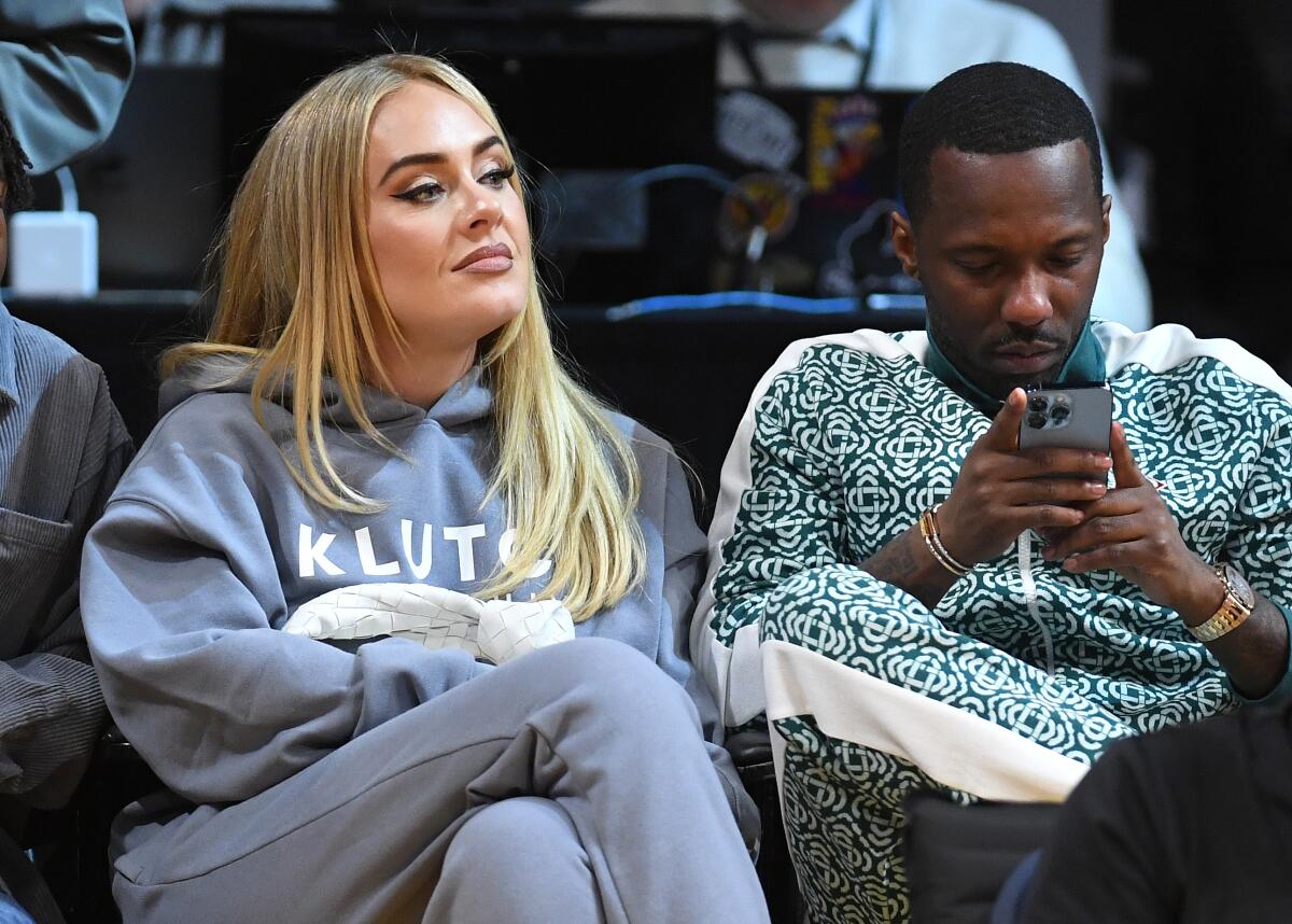 Adele sits with folded arms at a basketball game next to Rich Paul, who is looking at his phone
