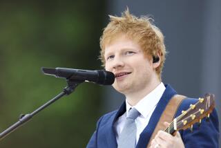 Ed Sheeran plays guitar and sings into a microphone while wearing a blue suit