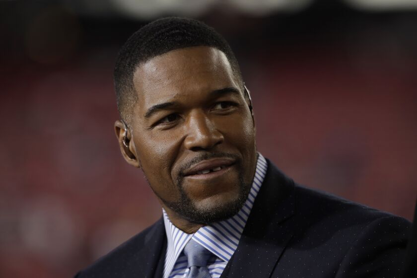 Michael Strahan wears a suit and tie in front of a reddish background