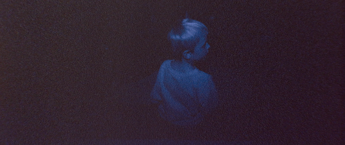 A young boy in the darkness, his back facing the camera, looking to the side