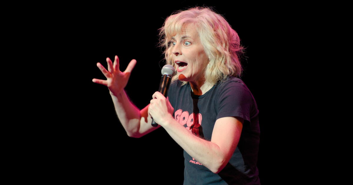 A woman in a black T-shirt with pink lettering gesturing with her hand while speaking into a microphone
