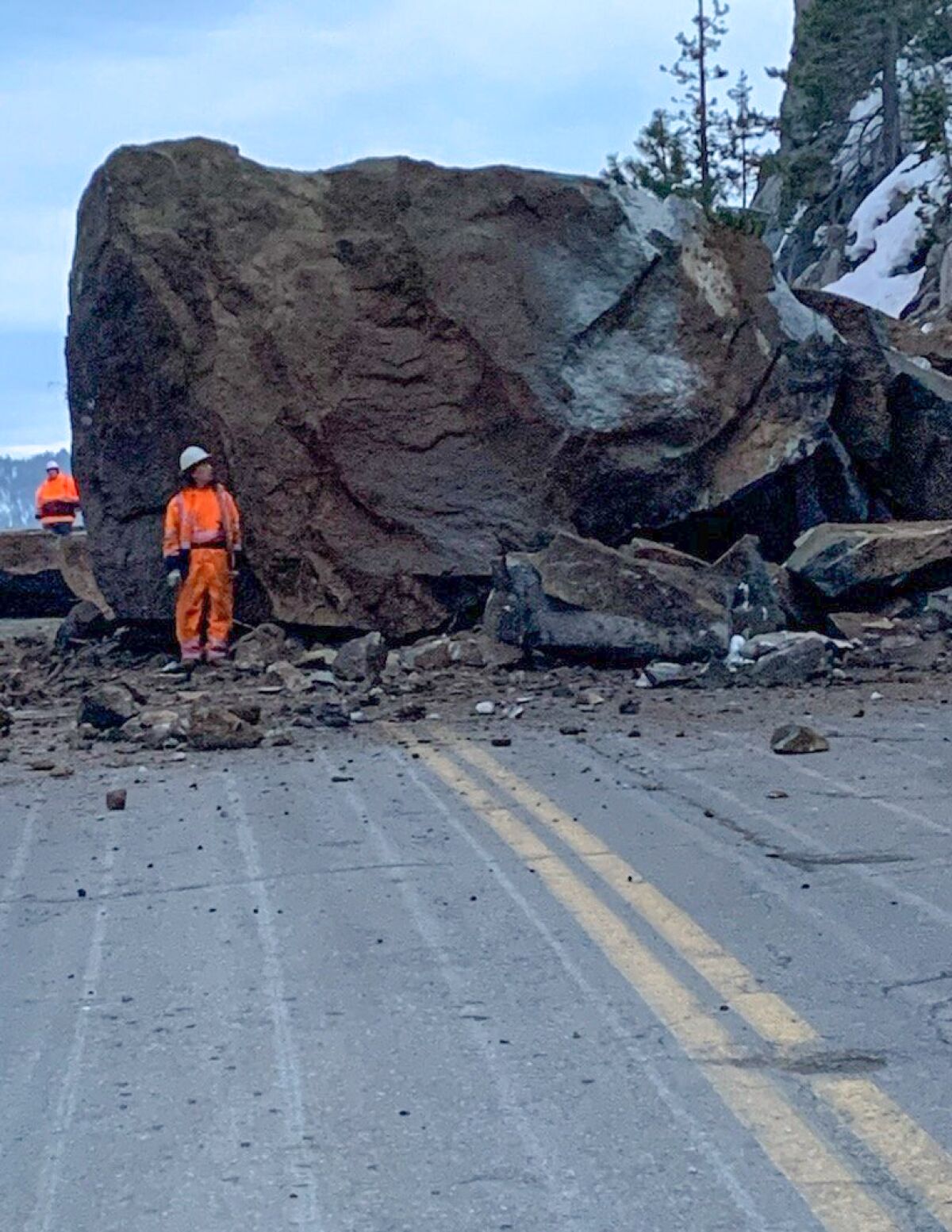 A boulder more than twice as high as a nearby worker blocks a two-lane highway