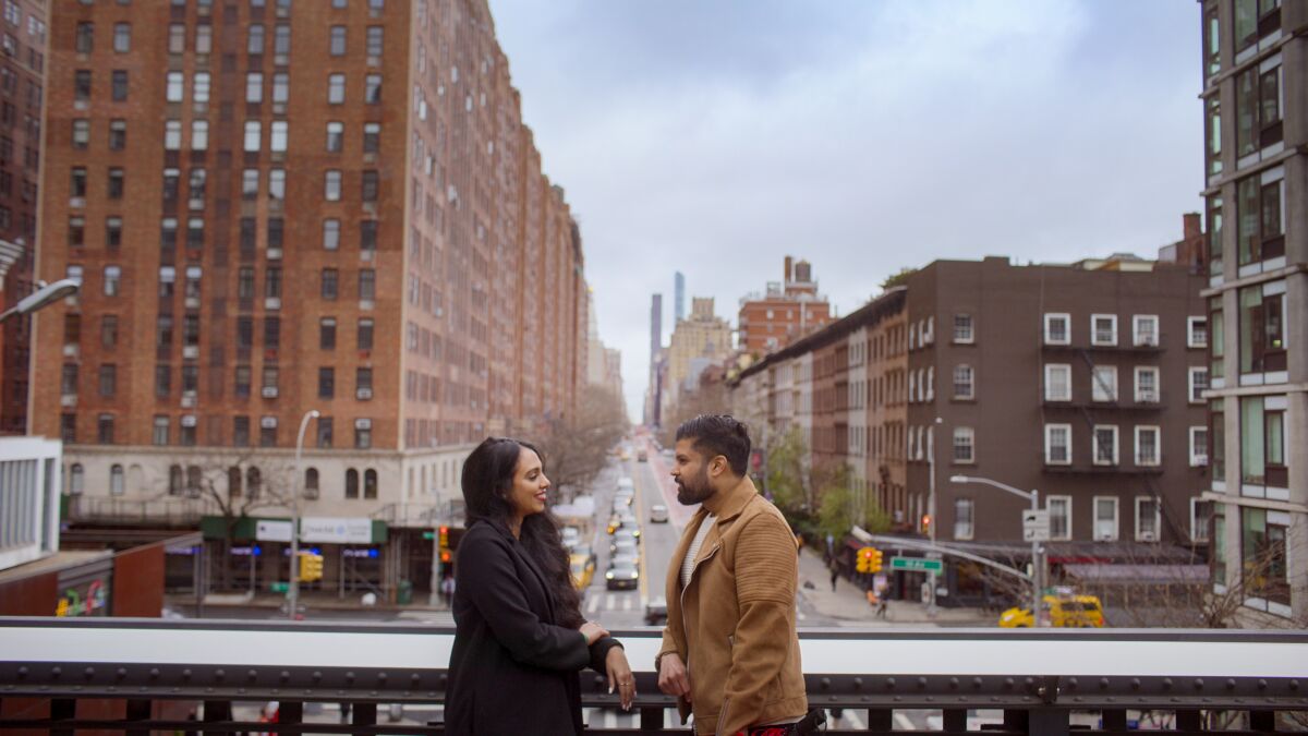 A couple look at one another with a city street stretching into the distance behind them.