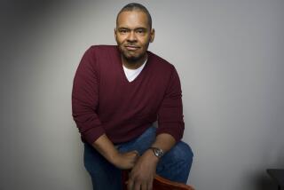 A Black man in a maroon sweater and white T-shirt and jeans posing with his left leg up on a chair against a grey background