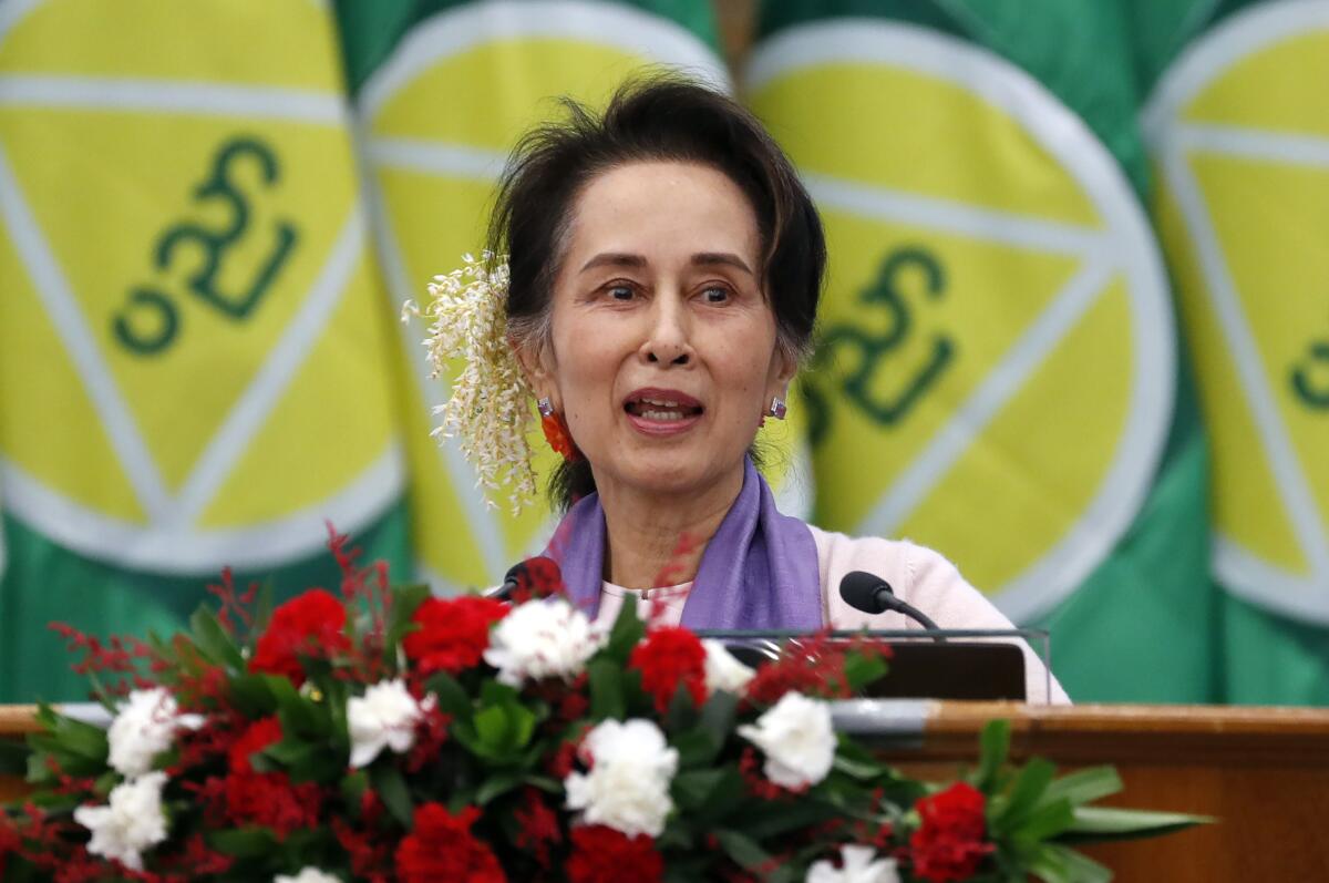 Aung San Suu Kyi delivers a speech with flowers in front of her and flags behind