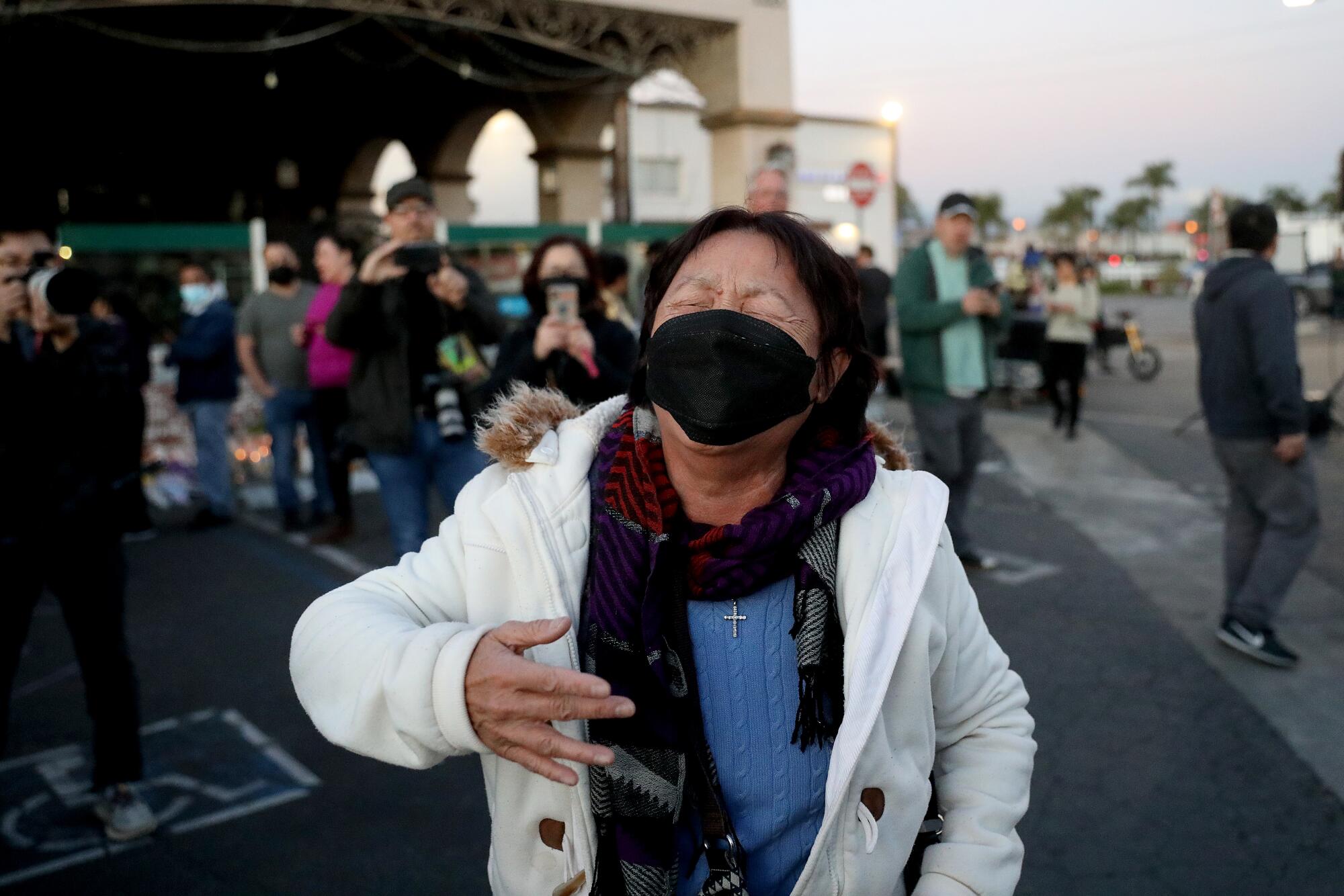 A woman wearing a white coat and a black face mask reacts and seemingly cries while gesturing with one arm.