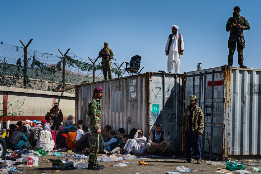 Taliban fighters stand on and by shipping containers as Afghans trying to reach the airport sit in a corner on the ground. 