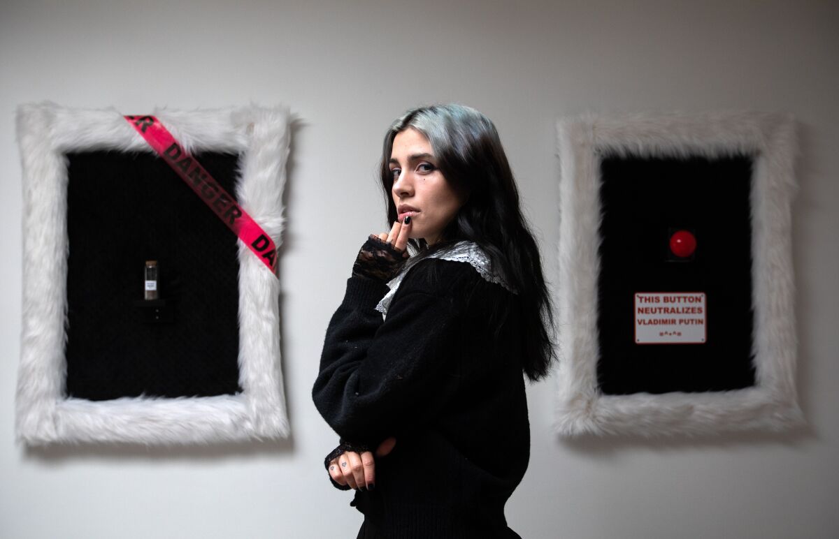 A woman stands in front of picture frames, one of which has a red button and reads, "This button neutralizes Vladimir Putin."