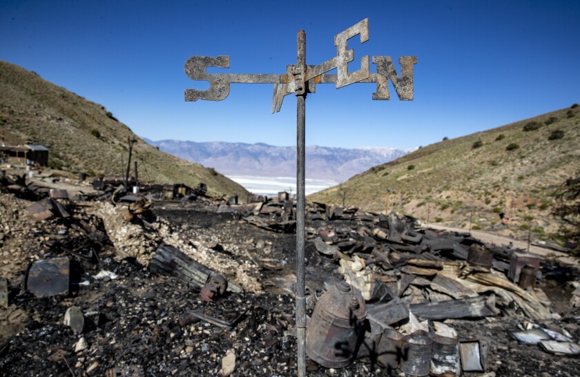 A weather vane blackened by fire stands among charred debris on a dry, brushy mountainside above a desert valley 