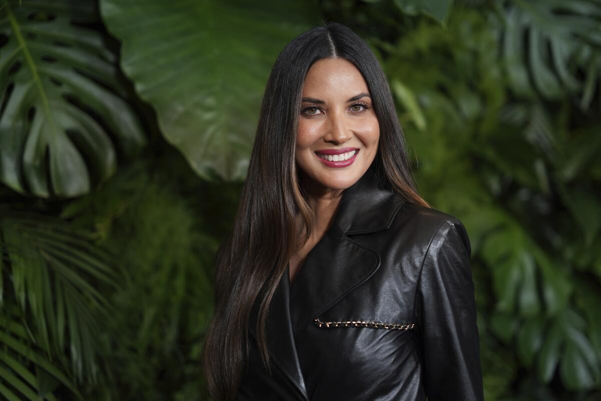 Olivia Munn smiling in a black leather jacket and long, straight dark hair in front of a wall of greenery