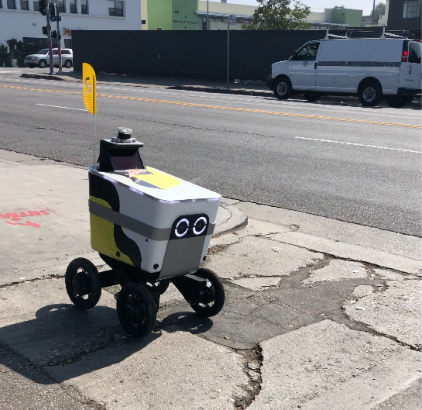A small robot resembling a cooler on wheels, with two illuminated circles for eyes, cruises along an L.A. sidewalk
