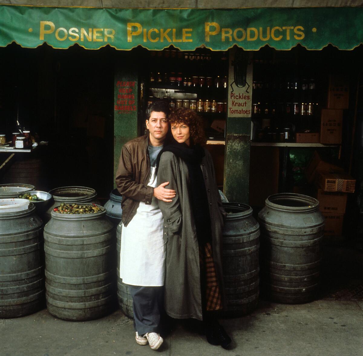 A man and a woman stand next to barrels in front of a pickle shop.